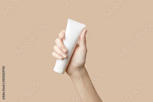 A womans hand holding a white cosmetic tube on a beige background. Care products