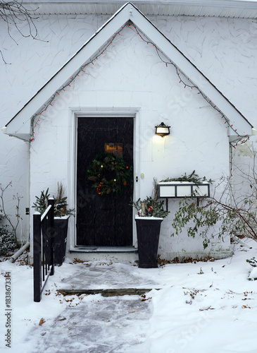 A small, steeply gabled white entryway with a black door, decorated with a wreath and flanked by seasonal planters, is visible in a snowy foreground in Ottawa