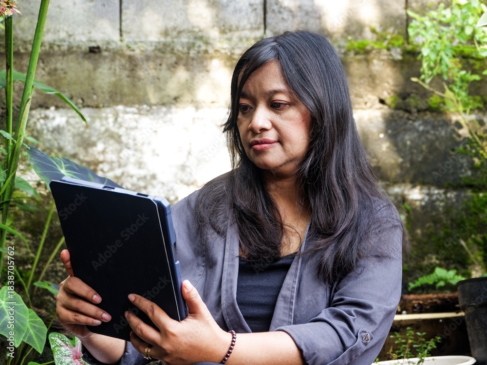 Fototapeta premium A professional woman intently using a digital tablet for remote work or creative design outdoors in a serene, tropical garden setting. Focus on technology and work-life balance.