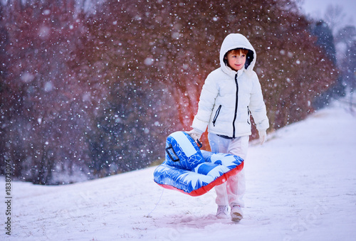 Happy Boy Walking Up Snowy Hill with Inflatable Sled – Winter Adventure in Falling Snow. Fun Seasonal Ride
