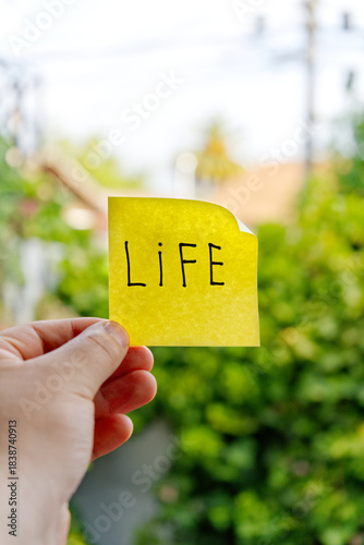 Photography A hand holds a yellow sticky note, boldly displaying the word LIFE, offering a message of inspiration and hope