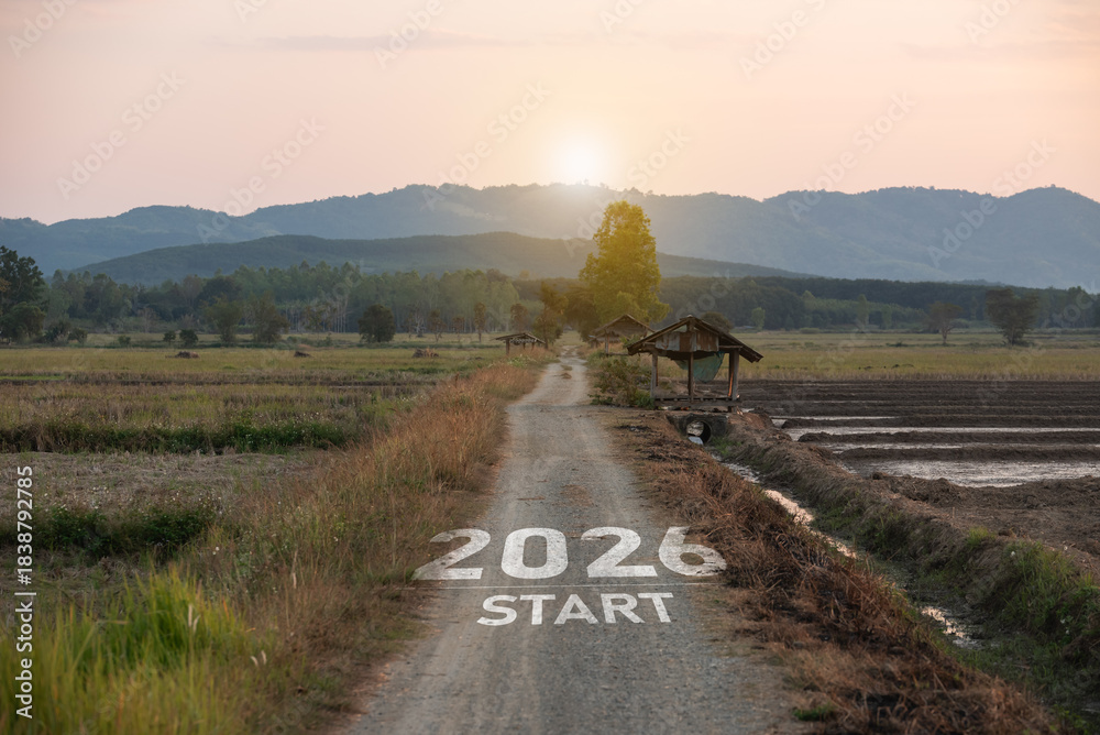 Naklejka premium New Year 2026. Scenic rural landscape featuring a dirt path marked with 2026 start, surrounded by fields, trees, and mountains at sunrise, ideal for vision and future themes.