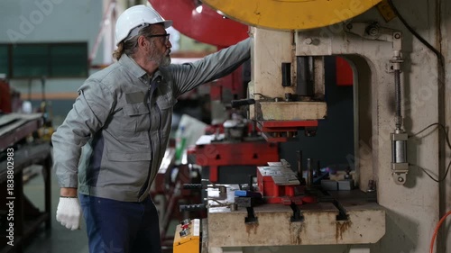 Senior industrial worker with grey beard and safety gloves operating a heavy red hydraulic press. He processes metal parts in a manufacturing factory workshop.