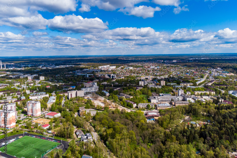 Fototapeta premium Aerial view of a sprawling city nestled among lush green spring foliage under a bright blue sky dotted with fluffy white clouds