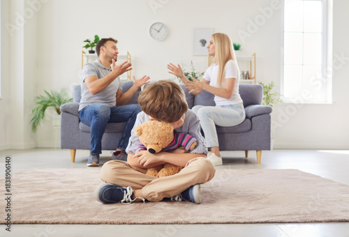 Foto Sad and anxious young boy sitting on floor, hugging his teddy bear, feeling powerless as his parents quarreling and arguing in background