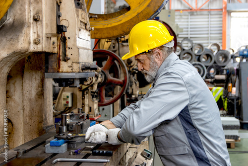 Senior male worker wearing safety hard hat and gloves while working with machine at manufacturing factory. Elderly engineer technician with helmet operating machinery in industrial factory site.