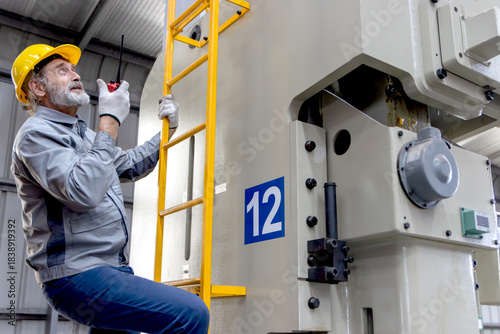 Senior worker wears safety hard hat, holds walkie talkie during climbs a ladder to inspect machine at manufacturing factory. Elderly engineer technician climbing up a ladder at industrial site.