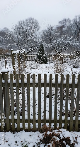 A snow-covered garden scene featuring a wooden picket fence in the foreground and frosted trees in the distance