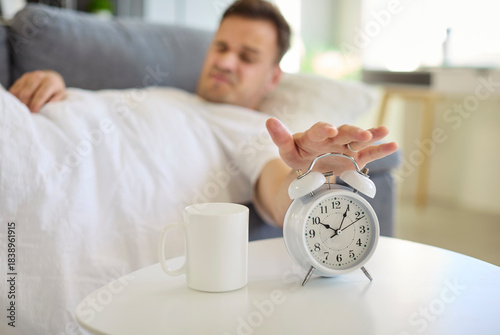Tired man reaching alarm clock in morning. In bed on a sofa, he fights waking and the impulse to press snooze during wakeup in a bedroom. It symbolizes reluctant waking and time management.
