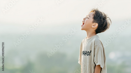 日本人の幼い少年が明るい屋外で、顔を空に向けて、大きな口を開けて大声で叫んでいる瞬間を捉えた横顔のポートレート。Tシャツには「YAHO!」と書かれており、喜び、自由、開放感の感情を表現。背景は霧がかったような柔らかい緑、崖の上