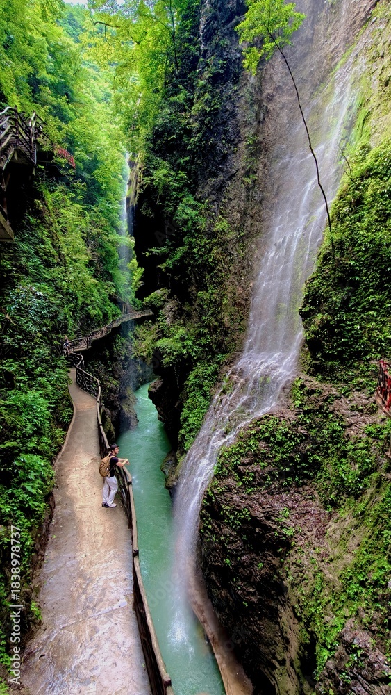 Fototapeta premium Male tourist enjoys Breathtaking gorge with a cascade waterfall, lush green cliffs, and a winding wooden boardwalk, showcasing the stunning natural beauty and accessible adventure of a Chinese canyon