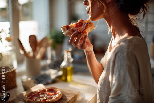 Young woman eating a warm pepperoni pizza slice in a cozy sunlit kitchen, holding the slice near her lips with rustic wooden utensils and olive oil nearby.