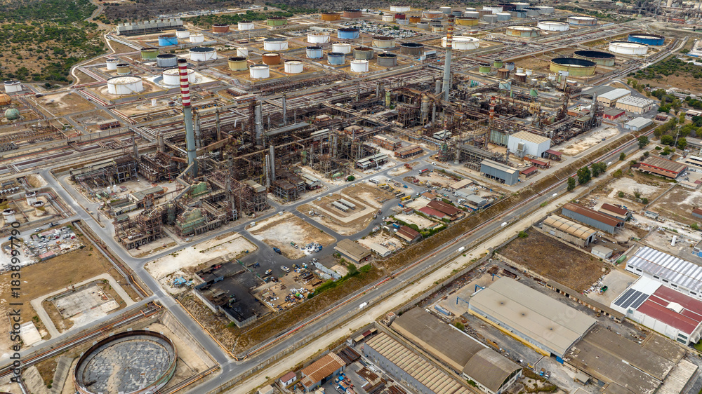 Fototapeta premium Aerial view of a large, complex oil and chemical refinery. The facility features a tall chimney, dense industrial piping and machinery, and a vast storage area of cylindrical tanks.