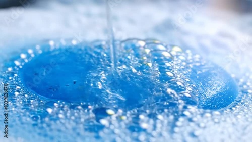 Close-up of clear water pouring into blue soapy liquid with many bubbles creating a relaxing, sudsy texture.