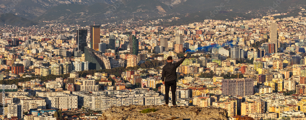 Obraz premium Man holding a blue smoke flare above Tirana’s skyline during golden sunset light.