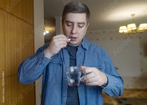Man taking dietary supplements at home, pouring pills from a bottle and drinking with water. Concept of daily vitamins, health care and wellness routine.