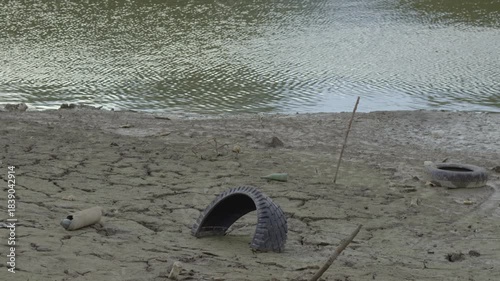 Dry cracked riverbank with low water level, discarded tires, bottle and other trash lying on muddy ground near rippled surface of reservoir.