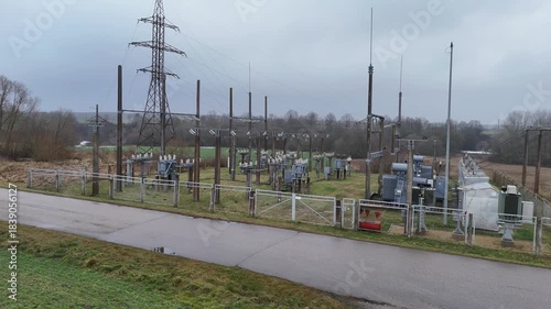 A fenced rural electrical substation surrounded by power lines, transformers, open agricultural fields and distant forest under an overcast sky, expressing industrial presence in natural countryside.