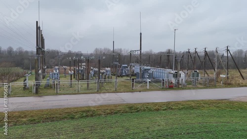 A fenced rural electrical substation surrounded by power lines, transformers, open agricultural fields and distant forest under an overcast sky, expressing industrial presence in natural countryside.