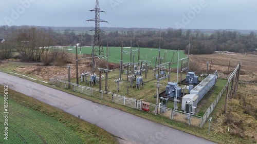 A fenced rural electrical substation surrounded by power lines, transformers, open agricultural fields and distant forest under an overcast sky, expressing industrial presence in natural countryside.