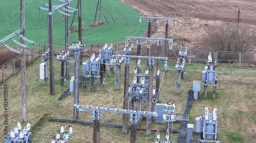 A fenced rural electrical substation surrounded by power lines, transformers, open agricultural fields and distant forest under an overcast sky, expressing industrial presence in natural countryside.