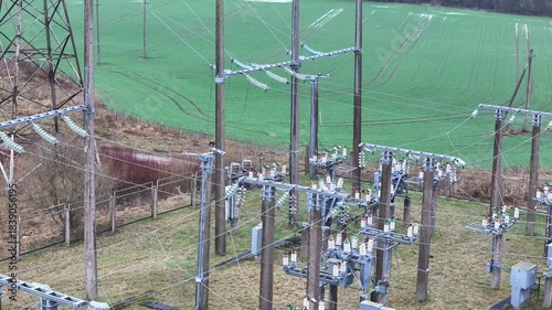 A fenced rural electrical substation surrounded by power lines, transformers, open agricultural fields and distant forest under an overcast sky, expressing industrial presence in natural countryside.