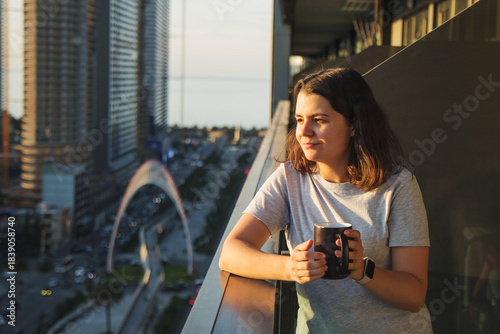 Woman enjoying a warm drink on a balcony during golden hour at sunrise or sunset, looking at the glowing city skyline and embracing a calm reflective moment concept lifestyle
