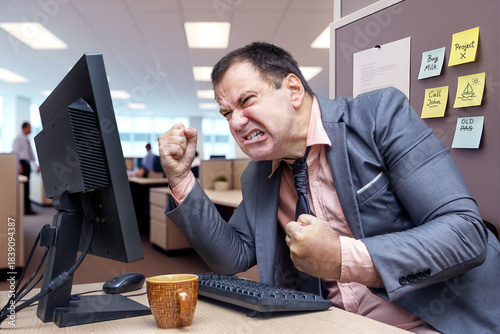 A stressed businessman in a gray suit clenches his fists at a computer screen in a busy office, showing frustration.