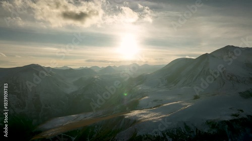 Aerial view of Majestic landscape of the Caucasus mountains at sunset. The sun shines brightly over snow-covered peaks, creating a dramatic lens flare and golden light over the valley.
