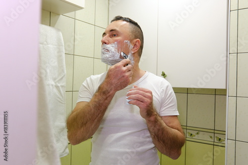 A man in the bathroom in front of a mirror applies shaving cream to his face. High quality 4k footage