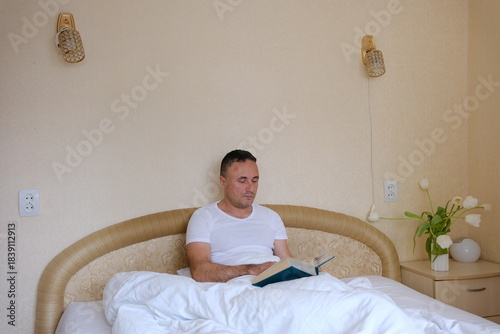 Man reading book in bedroom feeling serene and relaxed mood at night closeup. Engaged male reader absorbed in literature at lamp light. Calm guy enjoy hobby lying bed in tranquil home.