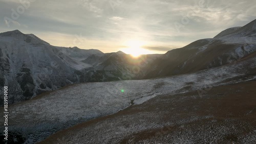 Aerial view of Majestic landscape of the Caucasus mountains at sunset. The sun shines brightly over snow-covered peaks, creating a dramatic lens flare and golden light over the valley.