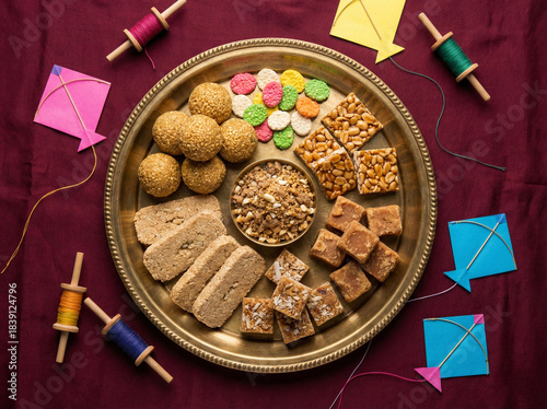 Brass plate filled with makarsankranti sweets and real kite festival decor arranged around the plate on a premium deep maroon matte background in clean top view festive composition