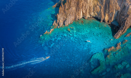 Aerial view of the turquoise waters gently lapping against the rugged, sun-kissed cliffs, boats cutting through the deep blue sea, Panarea, Sicily, Italy.