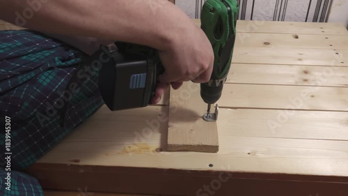 Male hands using green cordless drill to screw metal bracket into small wooden board placed on light wooden surface, person wearing plaid pants kneeling beside workpiece.