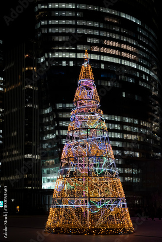 Paris, France - 12 08 2025: Christmas market. Panoramic view of the illuminated Christmas tree on the middle of the Parvis de la Défense and buildings behind at night