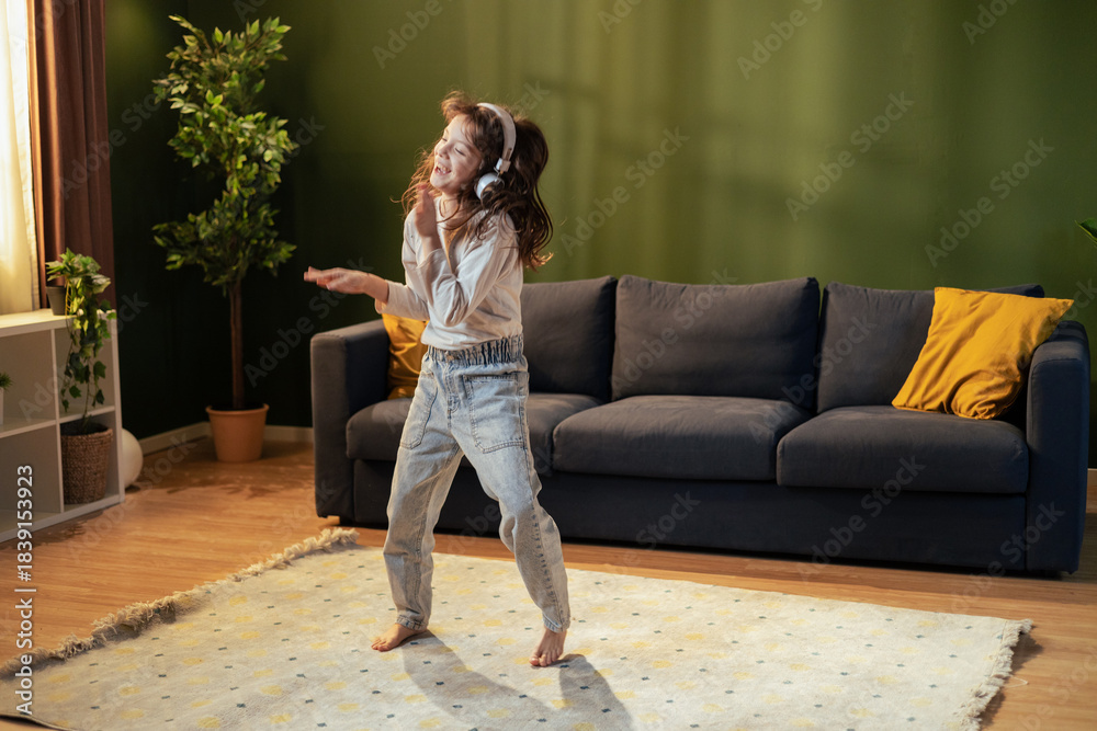 Naklejka premium Young girl dancing and listening to music with headphones in living room