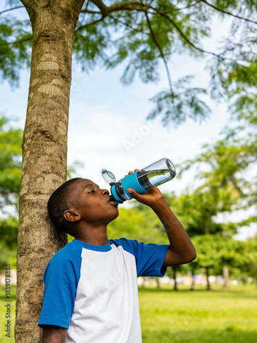 Black school-age boy in blue raglan tee drinking from clear reusable bottle with blue-grip flip-top, profile portrait leaning on tree in sunlit park — children's hydration, summer safety, eco-friendly