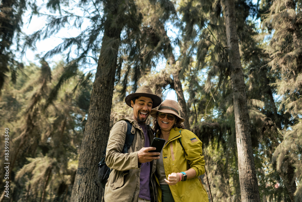 Obraz premium A couple of tourists look at their phone happily in the middle of the forest. They are wearing hats and hiking clothes