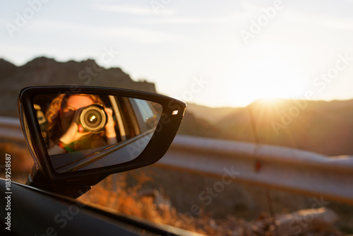 Reflection of a person taking a photo in a car side mirror during sunset.