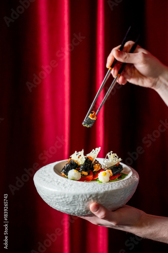 A close-up shot of hands using chopsticks to pick up a piece of gourmet, fine-dining food presented in a white bowl against a dramatic red curtain background