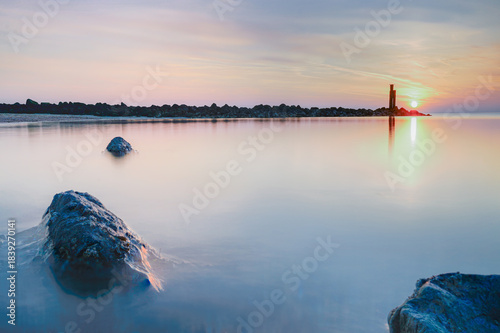 Prominent, textured rock in the shallow foreground, partially submerged in the receding water, reflecting the soft hues of the sky.