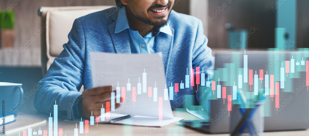 © Prostock-studio - An Indian entrepreneur reviews important documents in a contemporary office setting. He is focused on his laptop, browsing the internet and analyzing data with papers in hand.
