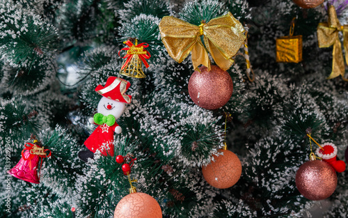 Festive Christmas ornaments hanging on a snowy pine tree. Glittery red and silver baubles, a gold and red bow, a snowman and a bell decorate the frosted green branches.