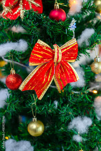 Festive Christmas ornaments hanging on a snowy pine tree. Glittery red and silver baubles, a gold and red bow, a snowman and a bell decorate the frosted green branches.