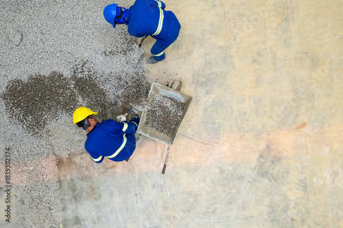 Workers on a construction site wearing yellow helmets and blue uniforms. Carrying a wheelbarrow of gravel.