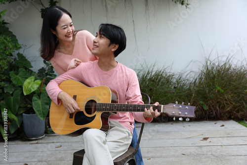 Asian young couple smiling in garden while man plays acoustic guitar and woman leans over his shoulder enjoying music and warm moment outdoors