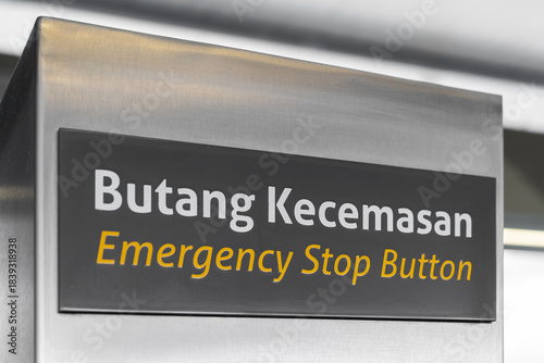 Industrial machine safety label in Malay and English. Emergency stop button communicates urgency, enabling quick shutdown and hazard prevention in critical situations.