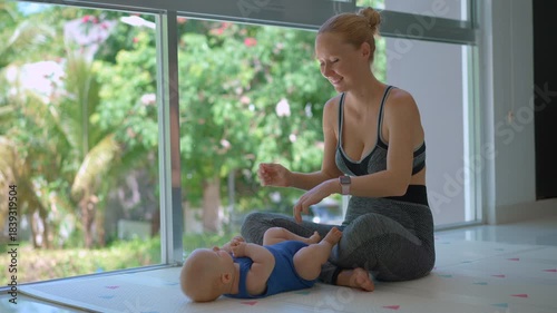 Young mother works out with her 3-month-old son at home by a big window with a lush tropical background.