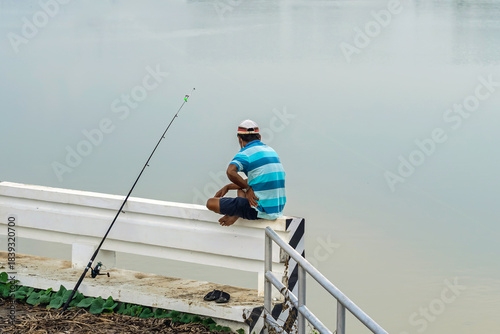 Elderly fisherman at side of river during sunset and catching fish with spinning. Senior fisherman with fishing rods on footbridge near the lake at summer. Mature man fishing on lake. Adult lifestyle.